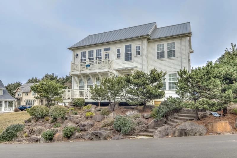 Two-story cream house in Lincoln City with a dark metal roof, white balcony, and stone steps