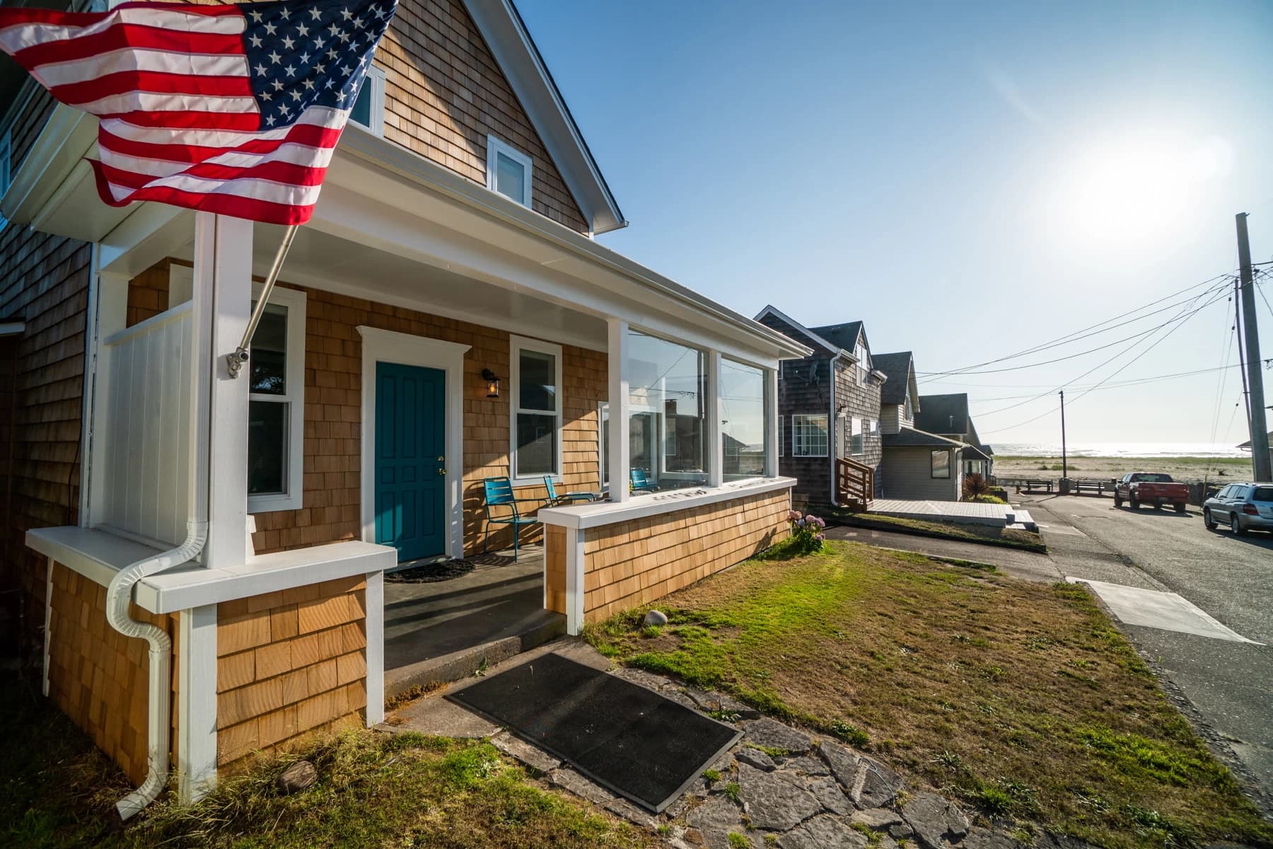 Cedar-shingle Seaside house with a teal door, American flag, and a street leading to the ocean