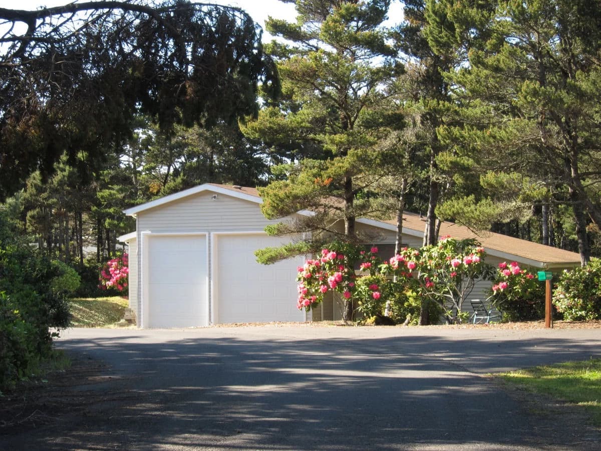 Beige house in Florence with two white garage doors, pines, and pink rhododendrons