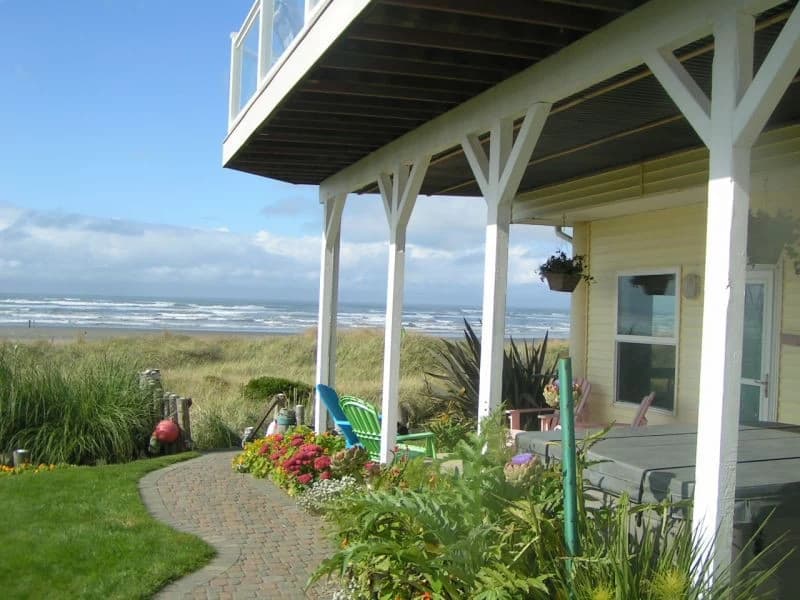 Yellow house in Moclips with a covered patio, gray hot tub, and ocean beyond dunes