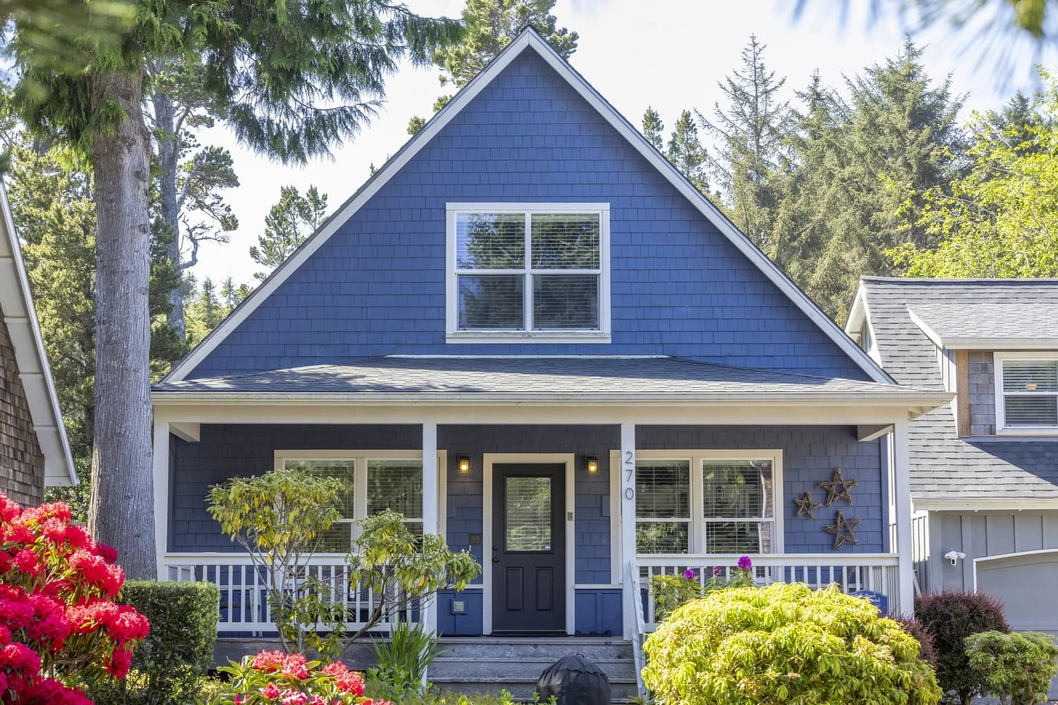 Dark blue two-story house in Lincoln Beach with a white porch and pink rhododendrons