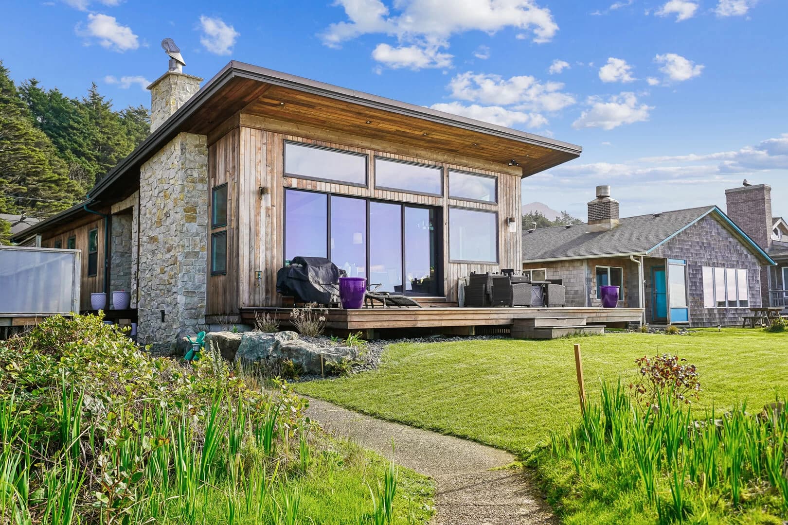 Wood-sided Arch Cape home with a stone chimney, glass walls, and furnished deck