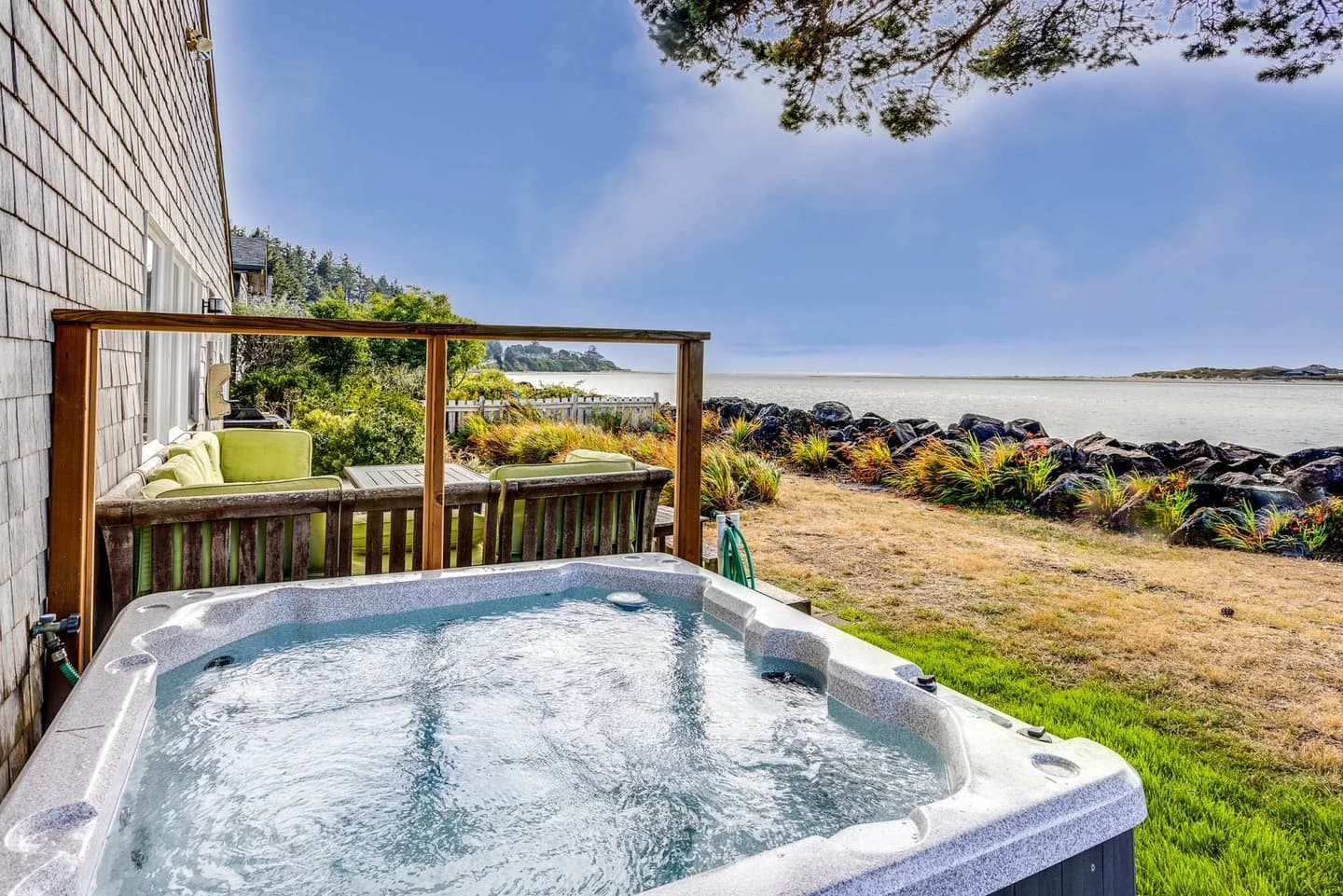 Hot tub by a cedar-shingle Waldport house with deck seating and rocky shoreline