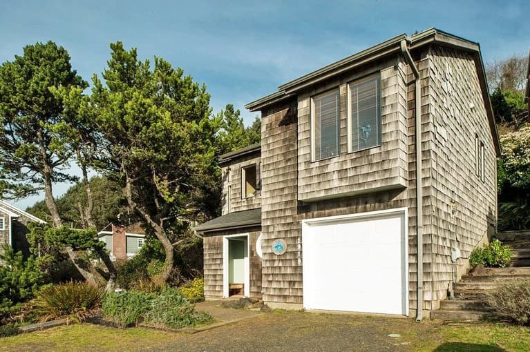 Gray cedar-shingle house in Lincoln City with a white garage door and evergreen trees