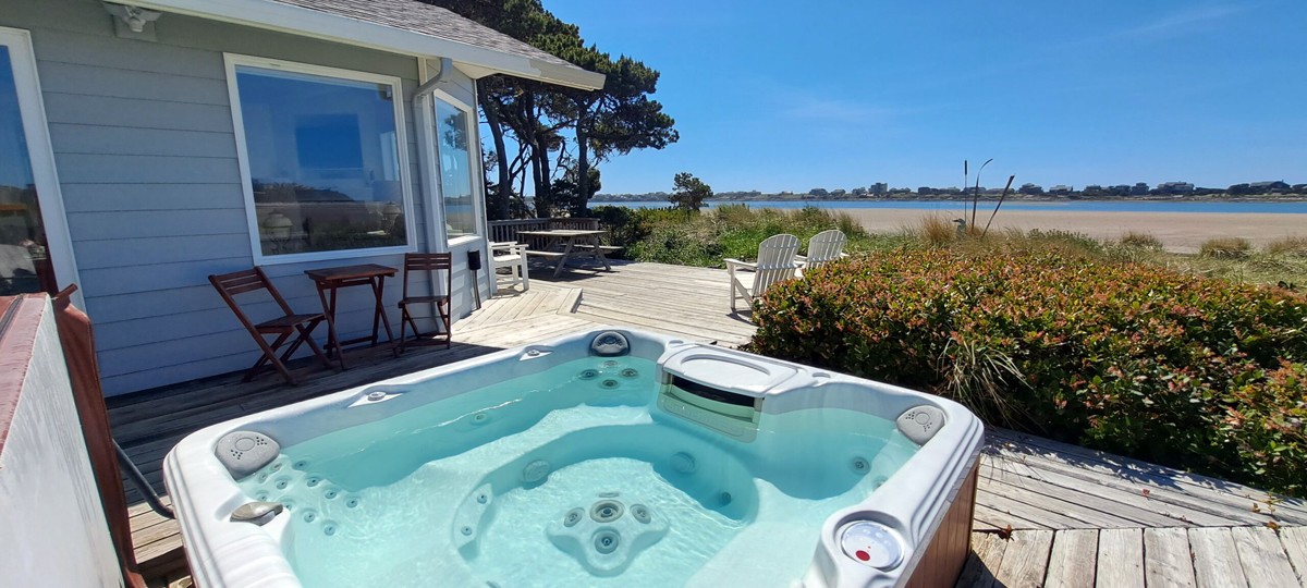 Inviting hot tub sitting on the side deck of a gray beach house with 2 white Adirondack chairs overlooking a sandy beach and waterfront view.
