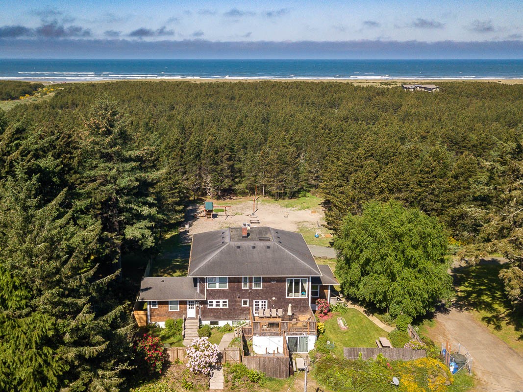 Aerial view of a 3-level large house surrounded by fir trees with the Pacific ocean in the distance.