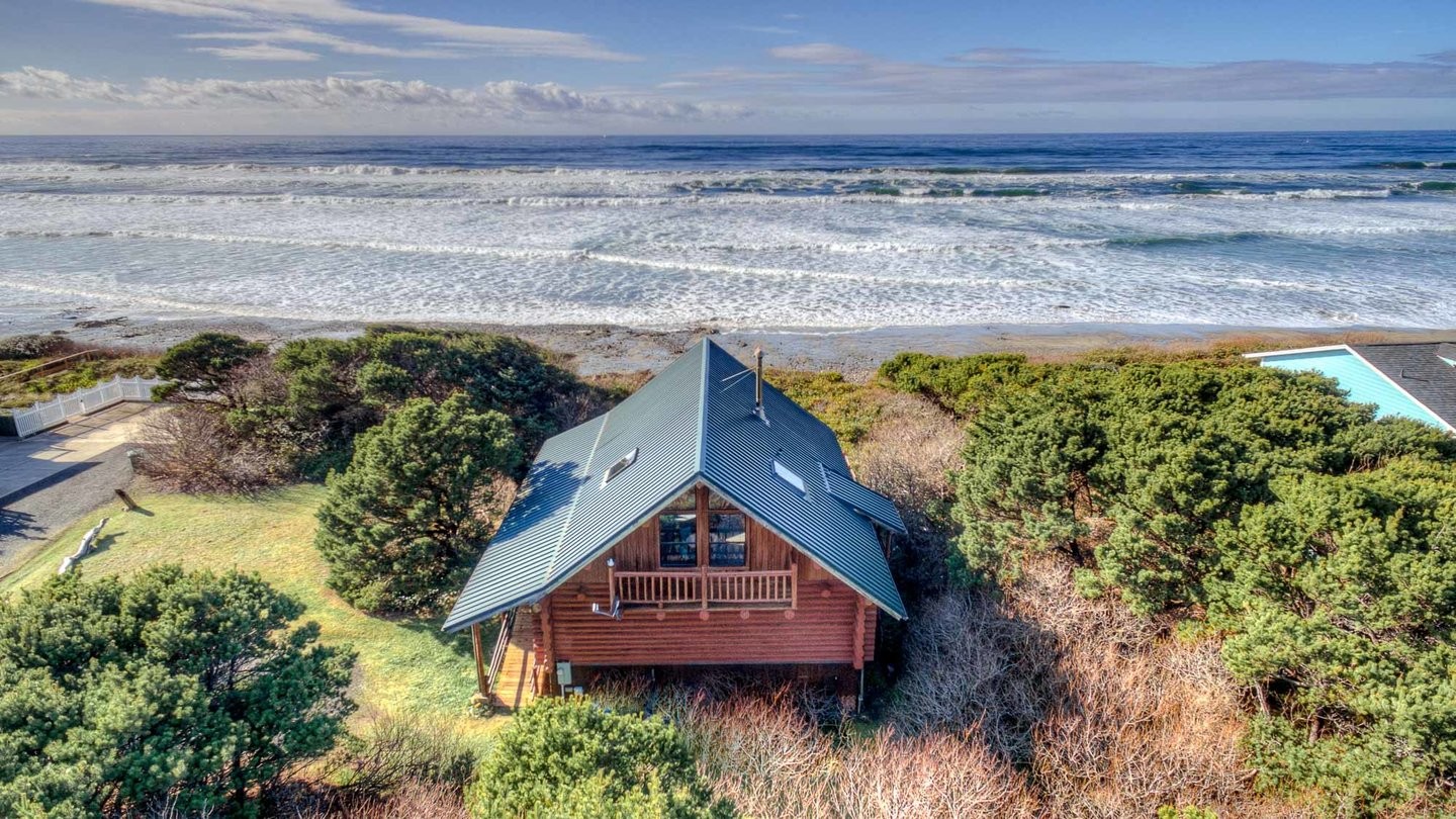 Aerial view of an oceanfront log house with a green metal roof in a natural tree lined setting.