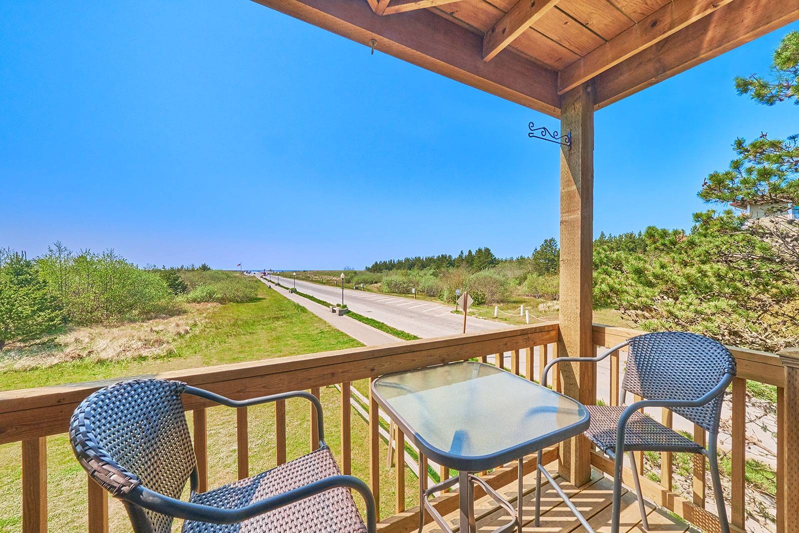 Corner deck of condo with table and chairs overlooking a paved approach to the beach on a blue sky day.