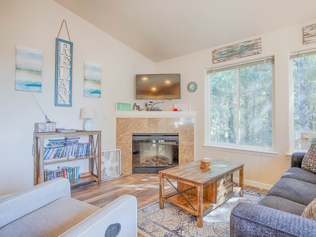 Sunlight shining on a small living room with a gas fireplace and TV above, a book shelf, coffee table, windows with a treed view, and a recliner and sofa.