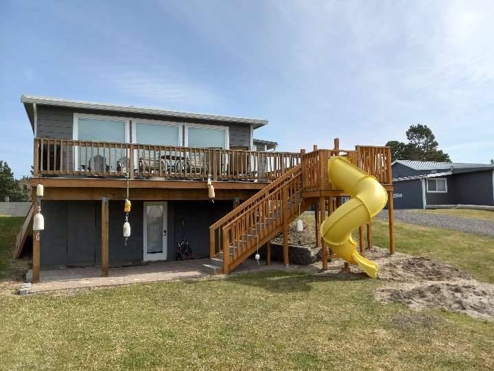 Two-level gray beach house with 3 large windows, a deck with stairs and a yellow tube slide off the top of the deck to the ground.