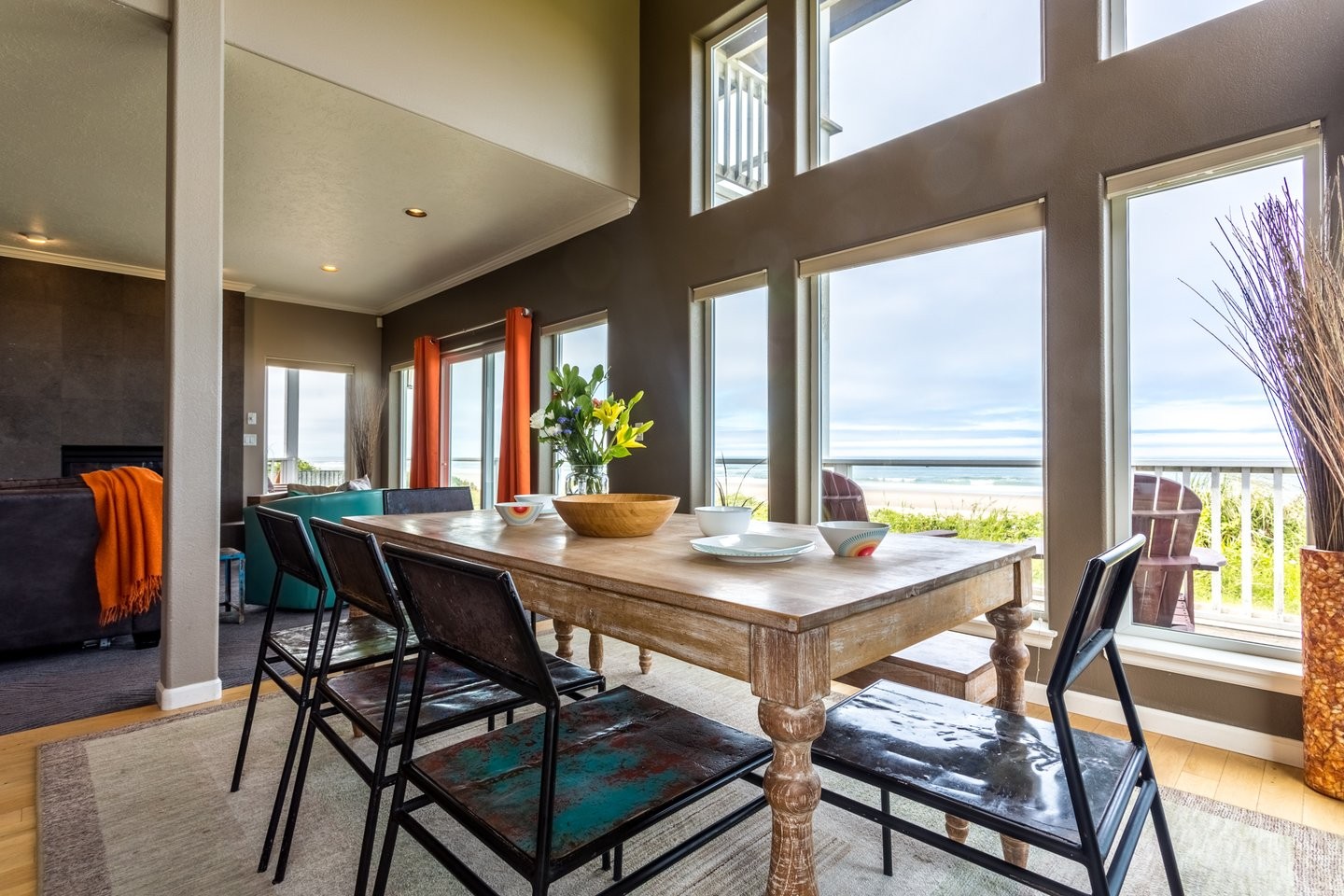 Luxurious open living room surrounded by ocean view windows, a large beautiful dining table, and colorful decor.